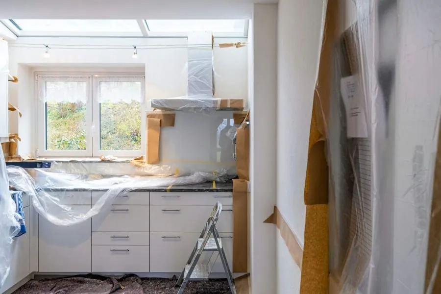 Kitchen under renovation, white cabinets, plastic sheeting, ladder, window, exposed walls.