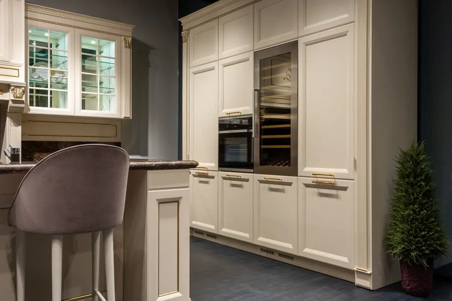 Modern kitchen with cream cabinets, built-in oven, wine fridge, and a barstool by a window.