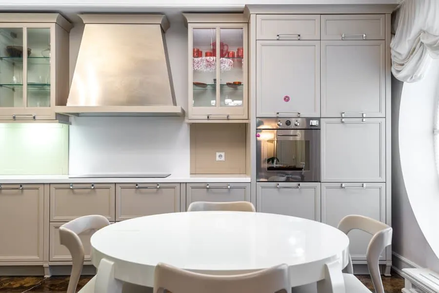 Bright modern kitchen with gray cabinets, white counters, built-in oven, and round white table by the window.