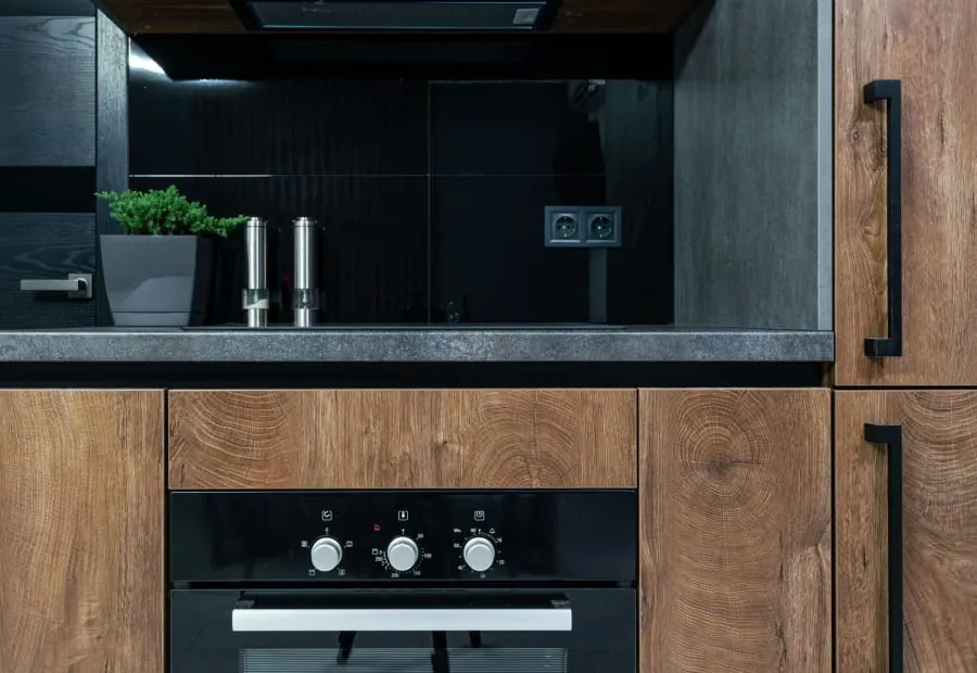 Modern kitchen with dark backsplash, wood cabinets, sink, and built-in oven under a countertop shelf