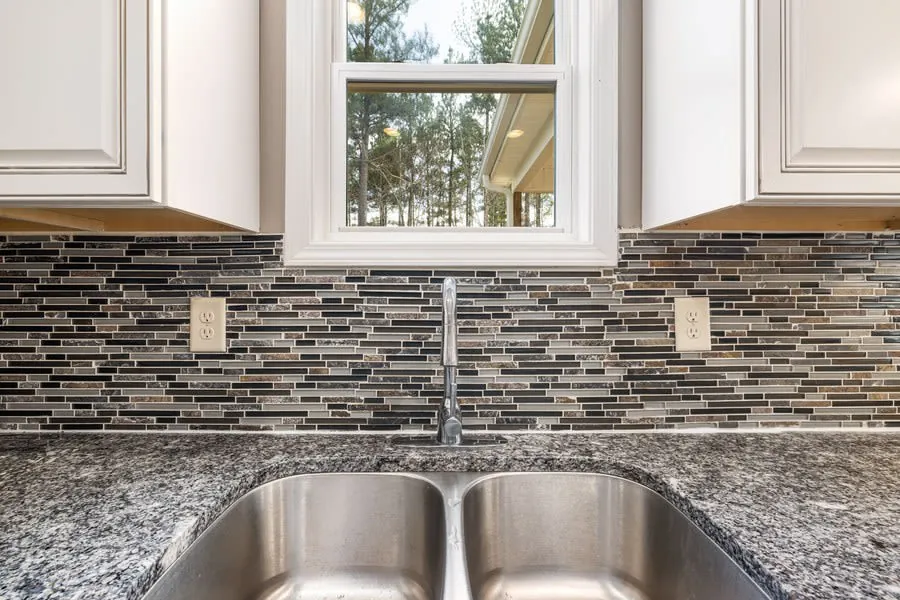 Kitchen sink beneath window with gray granite countertop and mosaic tile backsplash