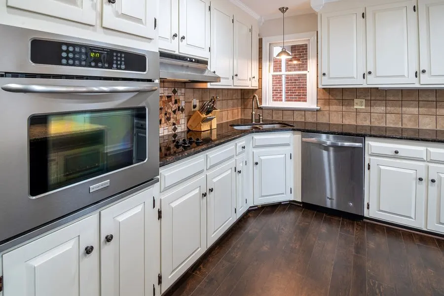 Bright white kitchen with stainless steel appliances, dark wood floor, and beige tile backsplash.