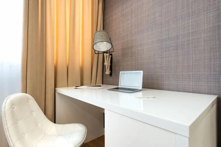 Compact home workspace with a laptop on a white desk beside a wall lamp
