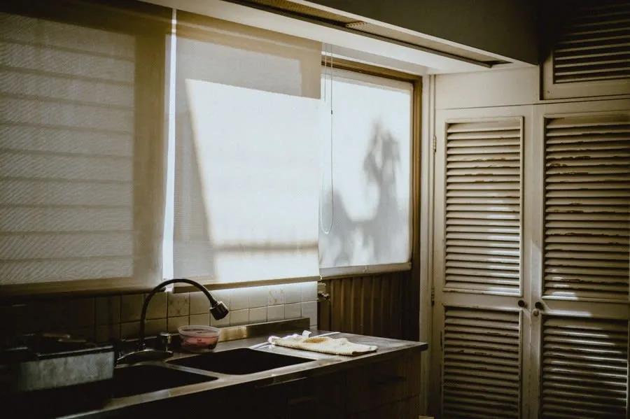 Older kitchen interior with worn units lit by daylight through window blinds