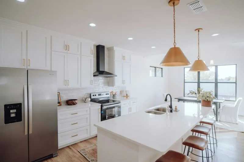 Modern white kitchen with island, stainless steel fridge, gold light fixtures, and a dining area.