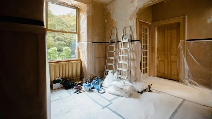 Room under renovation with ladders, tools, and plastic sheeting covering walls and floors.