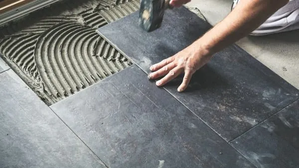 Person laying dark gray floor tiles with a hammer and trowel, indoors.