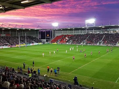 Rugby game in stadium under sunset sky; players on field, crowd in stands.